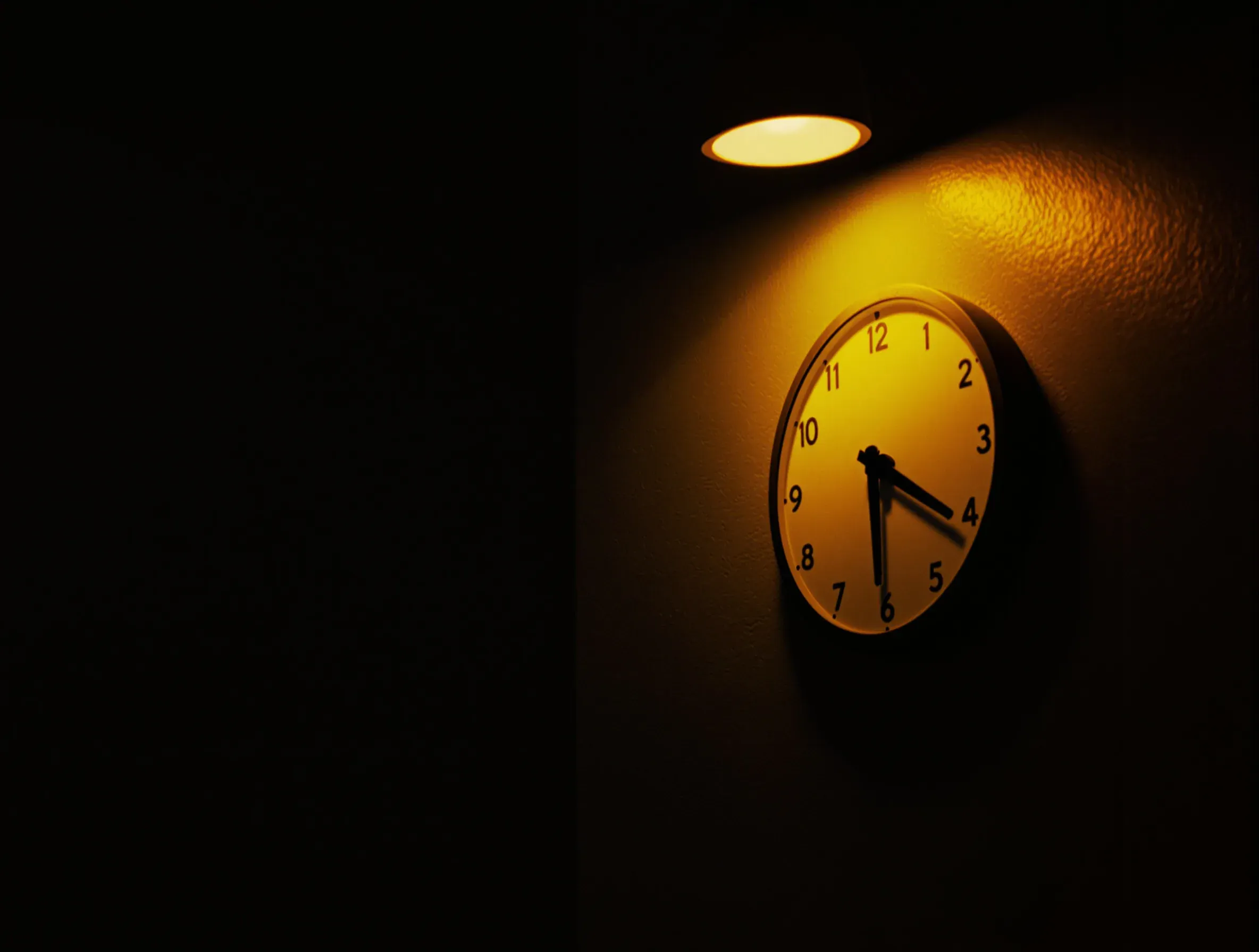 A plain wall clock lit by a single lamp in an otherwise dark room